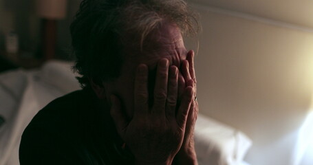 Old man sitting by the side of the bed at night rubbing face with hands