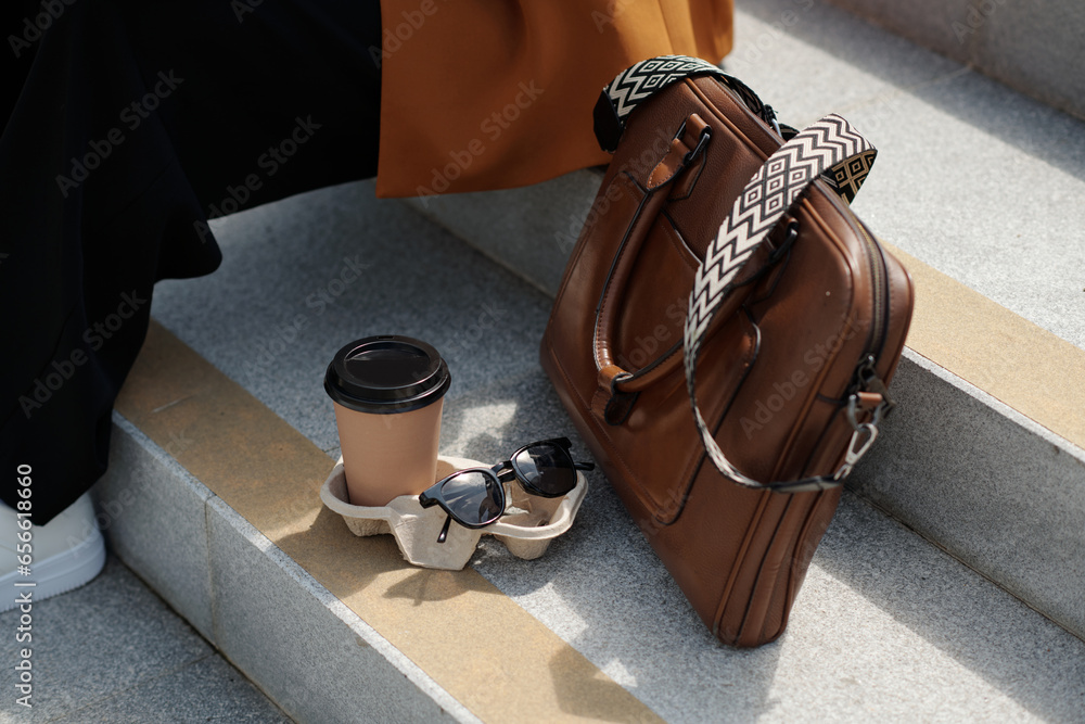 Canvas Prints Composition of sunglasses, cup of coffee and brown leather handbag on grey concrete stairs of staircase where young female employee sitting