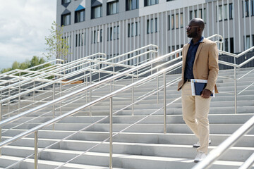 Young serious African American male solopreneur with financial documents and tablet in hand walking downstairs against building