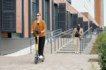 Young active businesswoman in eyeglasses and quiet luxury apparel riding electric scooter and moving along modern building of office center