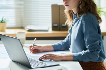 Side view of young serious female white collar worker in blue shirt sitting by desk in front of...