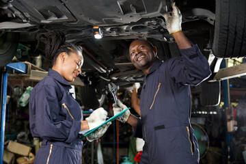 African mechanic workers fixing underneath car and talking about work in garage - obrazy, fototapety, plakaty