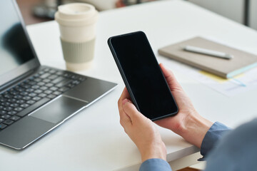 Empty screen of smartphone held by young businesswoman over desk with laptop, cup of coffee, notebook and pen during work in office