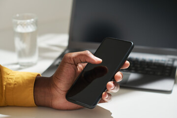 Hand of young African American office worker touching smartphone screen with thumb while sitting by desk and searching in the internet