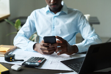 Cropped shot of young African American engineer texting in smartphone while sitting by desk with blueprints in front of laptop in office