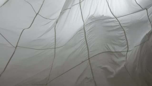 A huge parachute suspended from the ceiling in the pavilion sways in the wind, waves of white thin fabric, light shines through the fabric