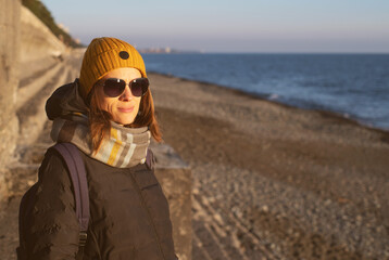 Portrait of a young woman in sunglasses, yellow knitted hat and scarf on the beach near the sea in the evening at sunset in winter, close-up, soft selective focus
