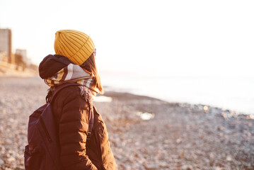 A young woman in a yellow knitted hat, scarf and with a backpack rests on a pebble beach by the sea...