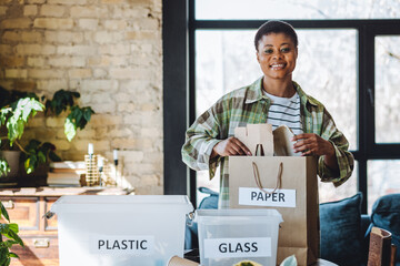 Young university student is managing waste sorting at home, smiling. Recyclable materials as paper,...