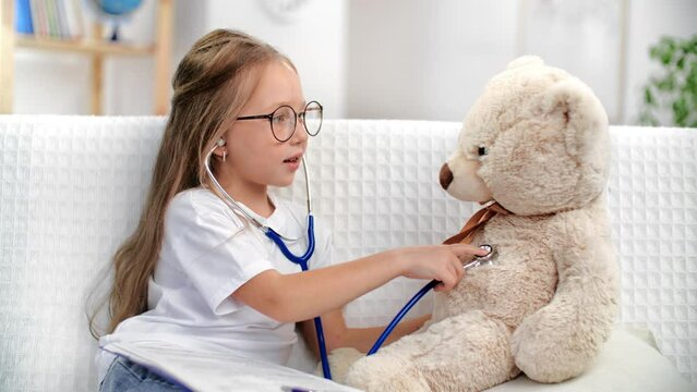 Confident blonde child girl playing imagine doctor talking bear toy patient use stethoscope at home