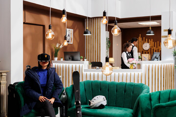 Female traveler wearing winter jacket occupies a sofa in lounge area and uses vr glasses to plan her skiing and snowboarding adventures. Woman using modern technology gadget in ski resort.