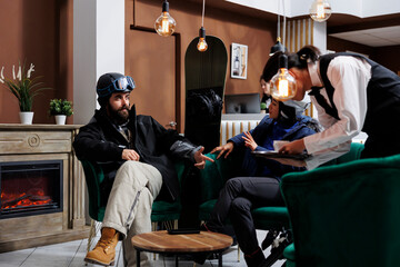 Couple in winter getaway sitting and conversing in ski resort lounge area while waiter serves...