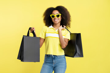 Stylish smiling curly haired African American woman wearing sunglasses holding black shopping bags isolated on yellow background, mockup. Shopping, black Friday concept