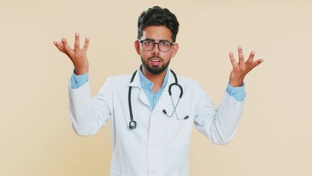What. Why. Confused Indian doctor cardiologist man raising hands in indignant expression asking reason of failure, demonstrating disbelief irritation by troubles. Apothecary guy on beige background