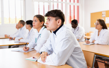 Young man medical student in white coat attedning classes in university, sitting at desk and listening to lecture.