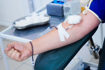 Donor sits in chair and donates blood from vein in medical clinic. Donor's hand with IV and needle...