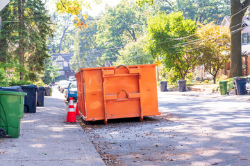 A roll-off construction dumpster parked on a residential street with traffic cones on a shady...