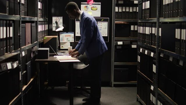 African American policeman carefully analyzes material, does records searches, and collects data from shelves and laptop. Male police officer standing, looking through case files on desk in office.