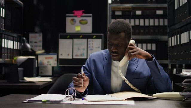 Focus on african american policeman answering phone call and taking important notes on case. In well-equipped office, expert male detective utilizes telephone to acquire evidence and analyze clues.