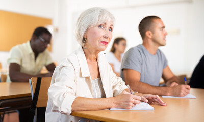 Senior woman sitting at desk in classroom while attending foreign language course with group of...