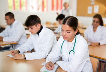 Young woman medical student in white coat attedning classes in university, sitting at desk and listening to lecture.