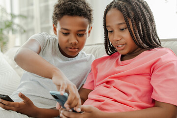 Smiling African American two kids sitting on the sofa and holding smartphones. Lifestyle, leisure, technology concept