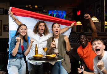 Group of excited young adults watching football championship in sports bar, supporting favorite team from Netherlands. People drinking beer with snacks at table and waving national Dutch flag..