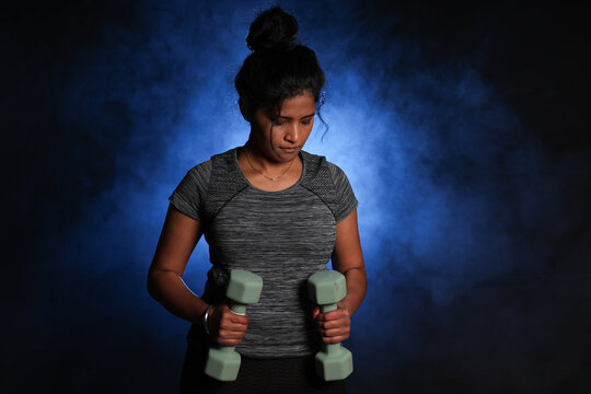 Young Woman Working Out with Dumbbells. She is dressed in active wear. Smokey background for added drama.