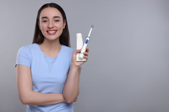 Happy young woman holding electric toothbrush and tube of toothpaste on light grey background, space for text