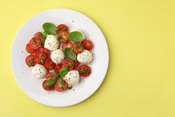 Tasty salad Caprese with tomatoes, mozzarella balls and basil on yellow background, top view. Space for text