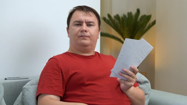 Plump man in red T-shirt sits on sofa sort mail, unpacking letters, carelessly crumpling bills, disdainfully throwing them away Unemployed debtor does not pay loans unpleasant information, invitations