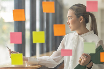 business asian girl in white shirt thinks, holds pen and clipboard, writing sticking adhesive notes on glass wall, making notes, standing over white background