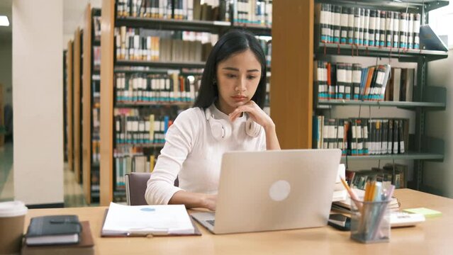 Asian undergraduate teen girl student study in library with laptop books doing online research for coursework, making notes for essay homework assignment, online education e-learning concept