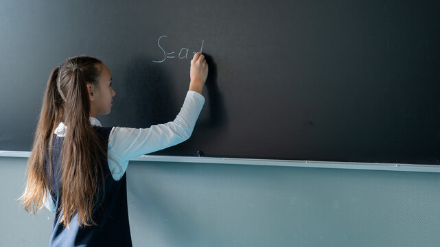 The schoolgirl answers at the lesson. Caucasian girl writes a formula on a blackboard.