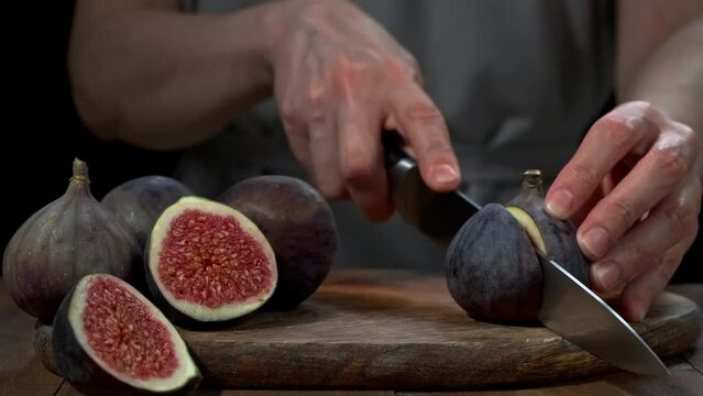 Women hands cut ripe fig in half with a knife on wooden cutting board. Close-up of whole and sliced fruits. Dark backdrop. Slow motion.