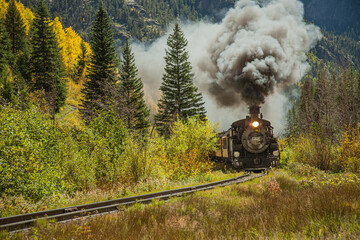 a steam engine train with smoke billowing from the smoke stack travels through the Colorado mountains an autumn day