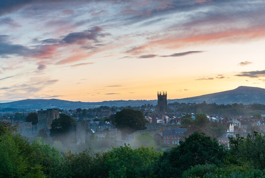 The Shropshire town of Ludlow with the Castle and Church and the rooftops of buildings prior to sunrise as mist forms in the foreground.