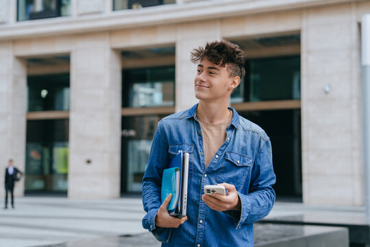 Satisfied European guy in jeans shirt holding notebook, folders and phone looks aside standing outside, passed exam. Smart Italian teen boy goes to university. Education, knowledge, youth.