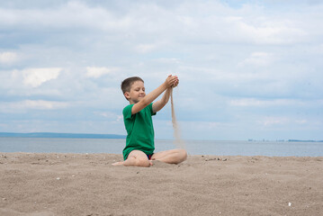 Light-colored child boy playing with sand on the beach against the background of water, blue sky, white clouds.