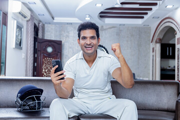 Overjoyed man in cricket dress celebrating win sitting on sofa at living room looking towards the mobile phone , Man enjoying watching cricket at home - obrazy, fototapety, plakaty