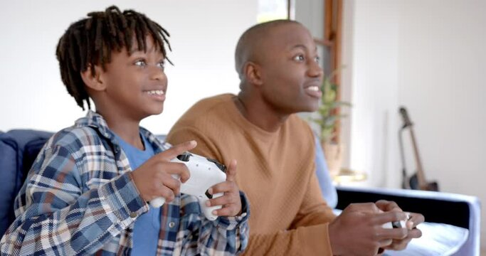 Happy african american father and son playing video games and fooling around at home, slow motion