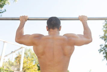 Back view of muscular sportsman doing pull ups on horizontal bar outdoors. Sports trainer on street workout exercising. Fit and athletic man having naked torso. 
