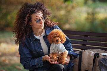 Portrait of beautiful curly haired woman holds small dog on her hands, while sitting in autumn park. Outdoor pet. Toy poodle dressed in clothes