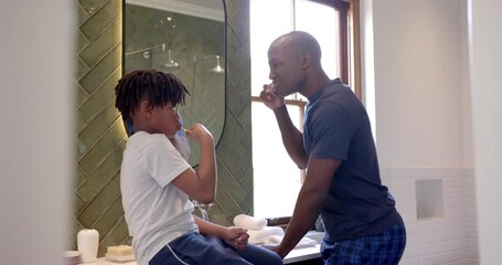 African american father and son brushing teeth in bathroom at home, slow motion
