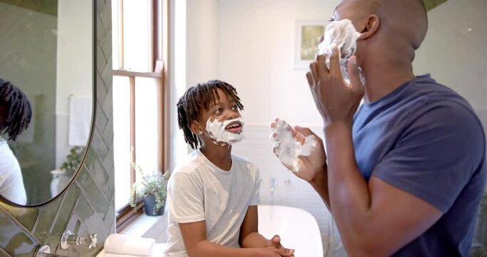 Happy african american father and son applying shaving foam on face in bathroom at home, slow motion