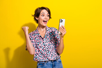 Portrait of astonished delighted girl use smart phone raise fist success achieve isolated on yellow color background