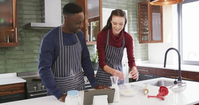 Happy diverse couple standing in kitchen, using tablet and preparing dough,slow motion