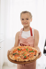 A little 8-year-old girl cook holds a delicious pizza while standing in a modern light kitchen.