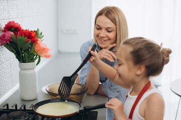 A young mother and her little daughter cook pancakes together for breakfast. Mother teaches daughter to cook.