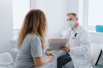 Fototapeta premium A woman at a doctor's appointment. A male doctor in a protective mask studies the medical record. Influenza pandemic, protective measures to prevent diseases.
